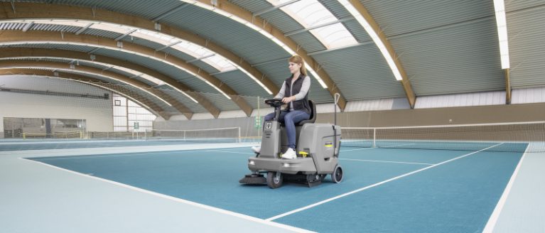 A woman cleaning the floor of an indoor tennis court with a ride-on carpet vacuum sweeper